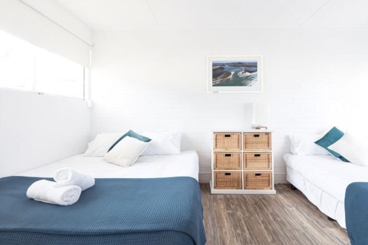 A hotel room with a bed, a small table, a nightstand, a window, and a wall-mounted picture.