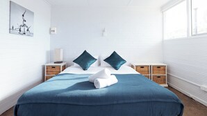 Een netjes opgemaakte hotelkamer met een groot bed, twee kussens, een blauwe deken en een witte handdoek. Er staan rieten manden op het nachtkastje en er hangt een schilderij aan de muur.