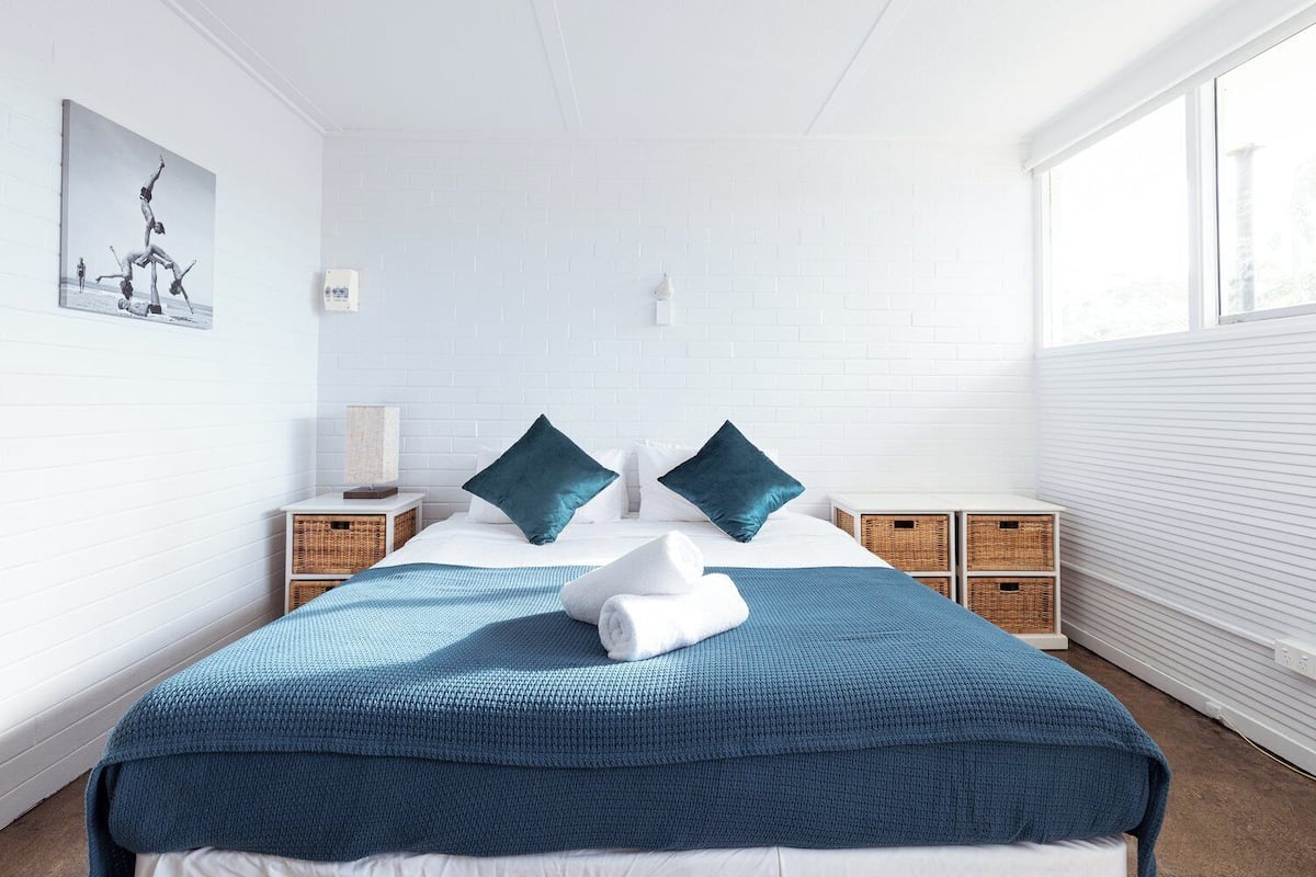 A neatly arranged hotel room with a large bed, two pillows, a blue blanket, and a white towel. There are wicker baskets on the nightstand and a painting on the wall.