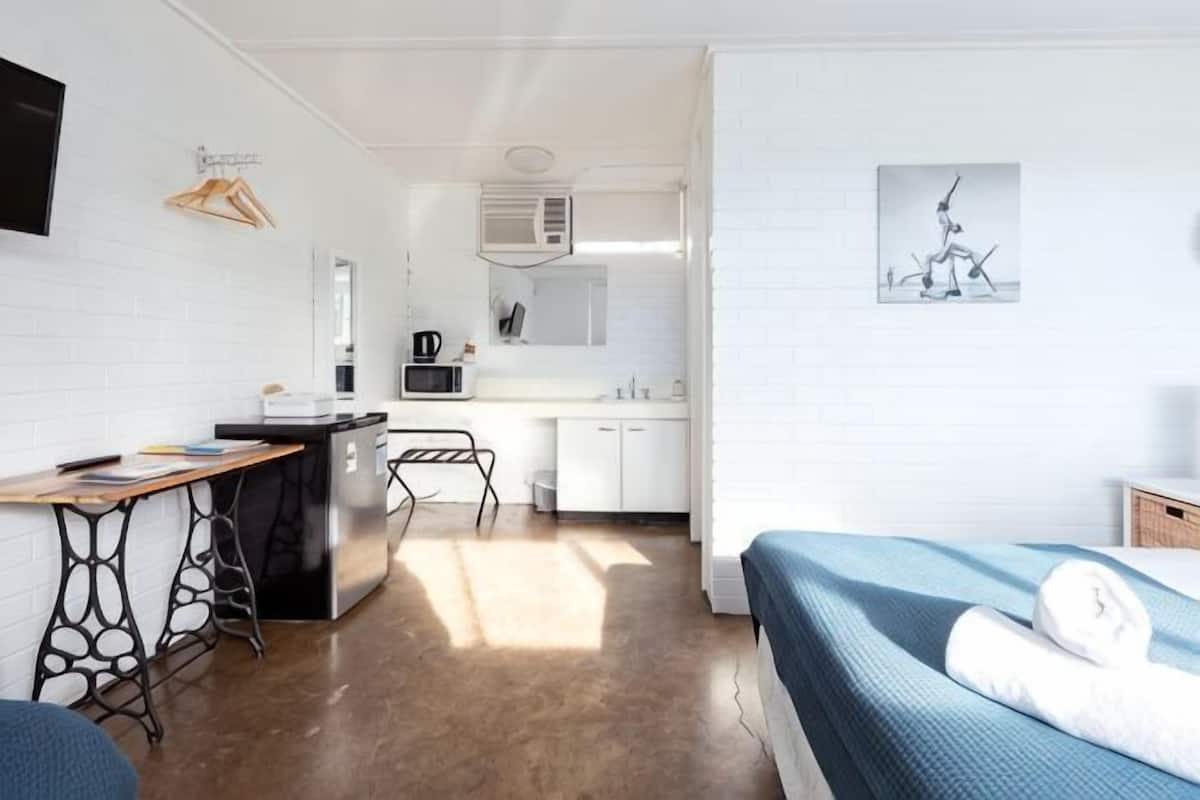 A compact hotel room with a kitchenette, a bed with a blue bedspread, a desk with a chair, and a wall-mounted TV.