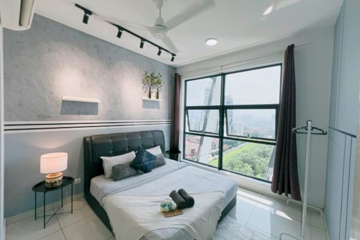 A modern bedroom with a large bed, a nightstand, a lamp, a window with a view of the city, and a ceiling fan.
