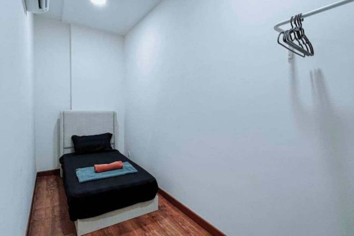 A small, narrow hotel room with a single bed, a white wall, and a wooden floor.