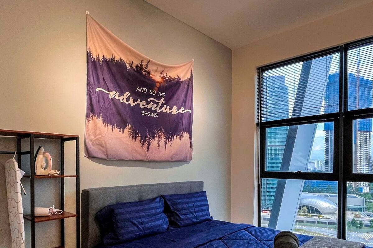 A bedroom with a city view, a bed with blue bedding, a grey headboard, a wardrobe, and a tapestry with a nature scene.