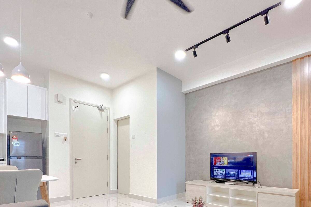A modern living room with a flat-screen TV, a dining area with a table and chairs, and a kitchen with white cabinets and a refrigerator.