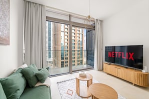 Een moderne woonkamer met een grote televisie waarop “NETFLIX” staat, een groene bank, een houten tv-meubel en een uitzicht op hoge gebouwen door grote ramen.