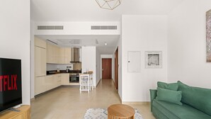 Un soggiorno moderno con un divano verde, un tavolino in legno e una TV che mostra 'NETFLIX'. Si intravede una cucina sullo sfondo, con mobili chiari e un'area pranzo.