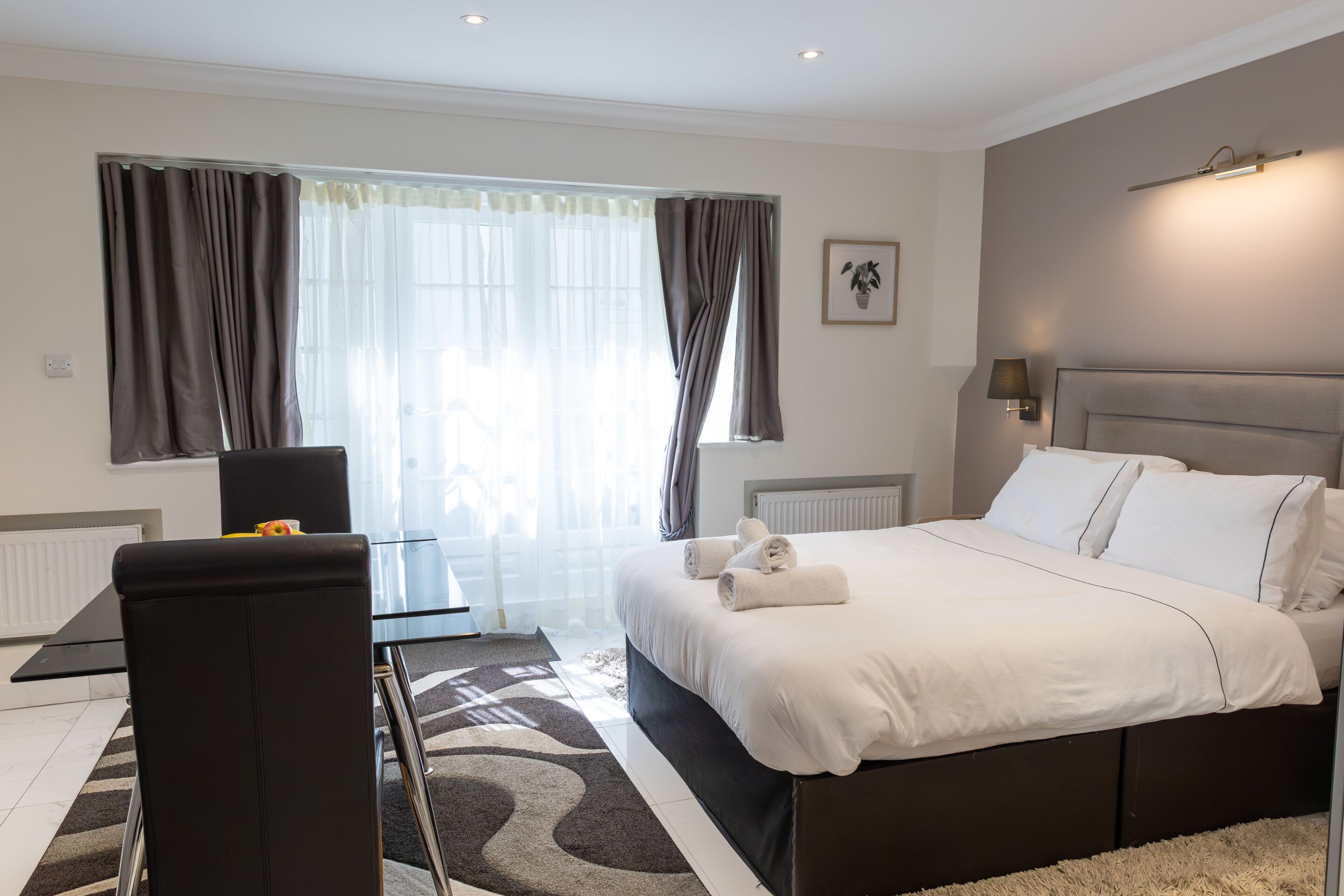 A modern hotel room with a large bed, a sitting area with a table and chairs, and a view of the outdoors through the window.