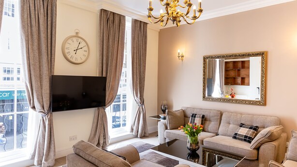 Un salon comprenant un canapé, des fauteuils, une table basse, un téléviseur, un lustre et une grande fenêtre avec des rideaux.