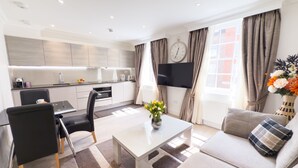 Un salon moderne avec un canapé, une table basse et un coin repas comprenant une table et des chaises. Un téléviseur à écran plat est fixé au mur, et une horloge est visible.