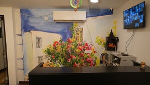 Un mostrador de recepción con un mural colorido, un equipo de aire acondicionado, un televisor y un pequeño santuario.