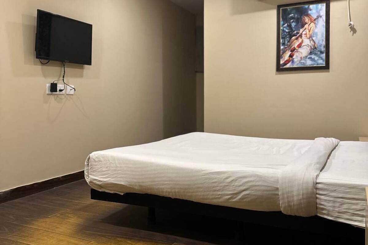 A hotel room with a bed, a ceiling fan, an air conditioning unit, a mounted TV, and a painting on the wall.
