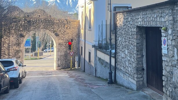 Una calle con un arco de piedra, un edificio de fachada de piedra y una cordillera al fondo.