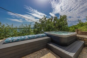 A hot tub with a wooden deck and steps, surrounded by greenery and a clear sky.