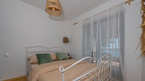 Ein Schlafzimmer mit einem Bett, einer Hängelampe, einem Fenster mit durchsichtigen Vorhängen und einer kleinen Wanddekoration.