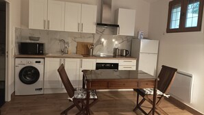 A compact kitchen with white cabinets, a microwave, a washing machine, a dining table with two chairs, and a window with blinds.