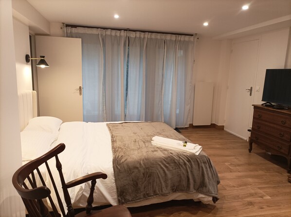 Un dormitorio con cama, una silla de madera, cómoda, televisor y ventana con cortinas.