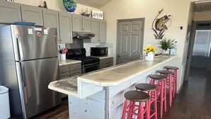 A modern kitchen with a stainless steel refrigerator, a microwave, and a stove. There are four red bar stools around a central island. - Pompano - Beachfront - Island Realty (Grand Isle)