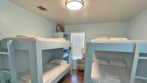 A room with two bunk beds, wooden flooring, a window with blinds, and a small table with a lamp. - Pompano - Beachfront - Island Realty (Grand Isle)