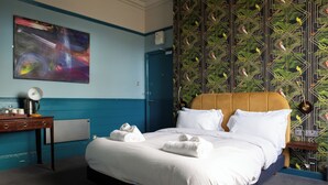 A neatly made bed with white linens and rolled towels, a wooden nightstand with a lamp, and a wall with a colorful abstract painting and a patterned wallpaper. - The Ship Rooms (London)