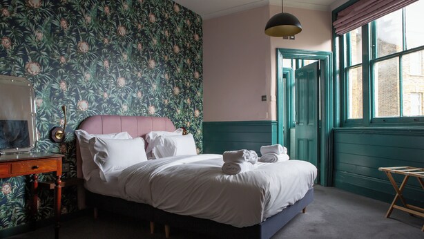 A bedroom with a floral wallpaper, a bed with white linens, a wooden nightstand, and a window with a view of the outside. - The Ship Rooms (London)