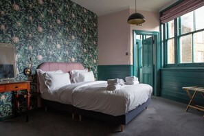 A bedroom with a floral wallpaper, a bed with white linens, a wooden nightstand, and a window with a view of the outside. - The Ship Rooms (London)
