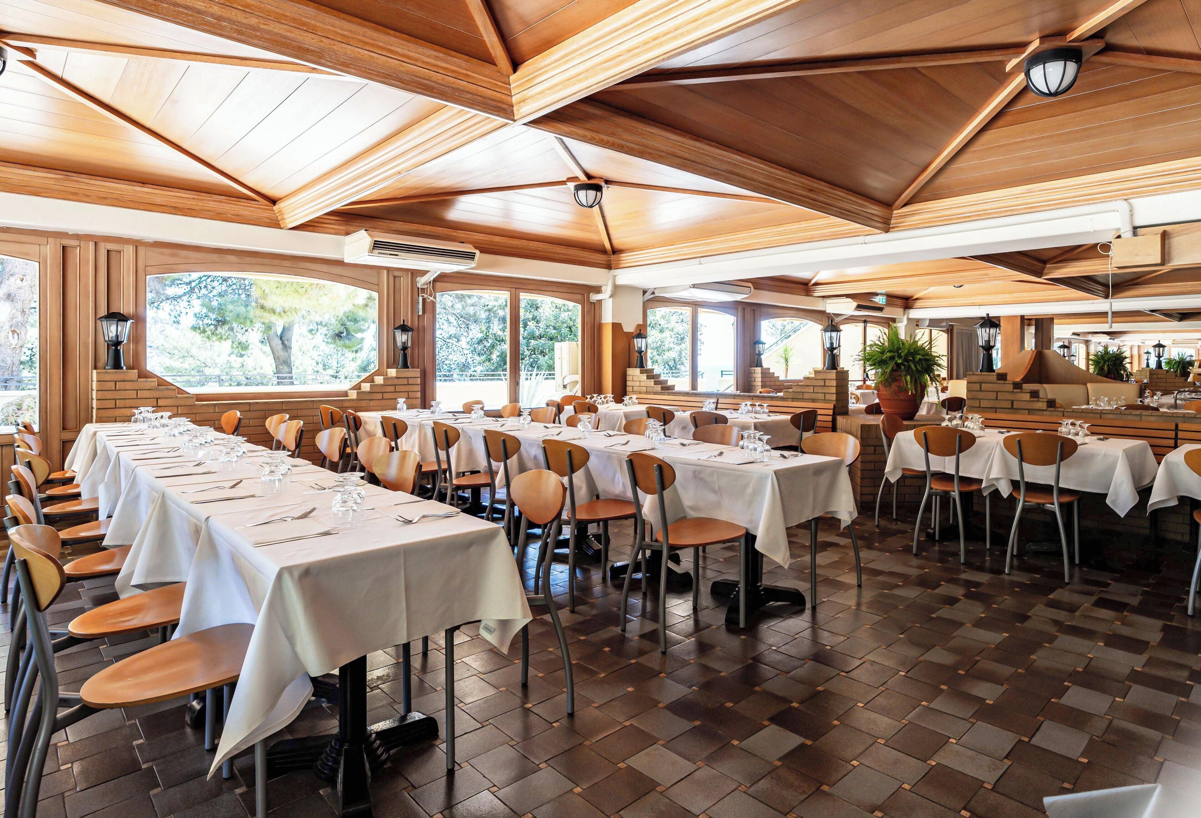 Ein Restaurant mit mehreren Tischen und Stühlen, einer Holzbalkendecke und großen Fenstern.