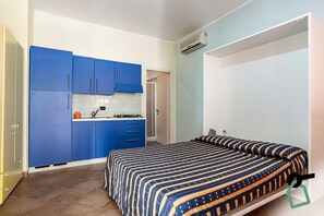 A compact room with a bed, a kitchenette with blue cabinets, and an air conditioning unit. - Hotiday Apartment Collection-Pugnochiuso (Vieste)