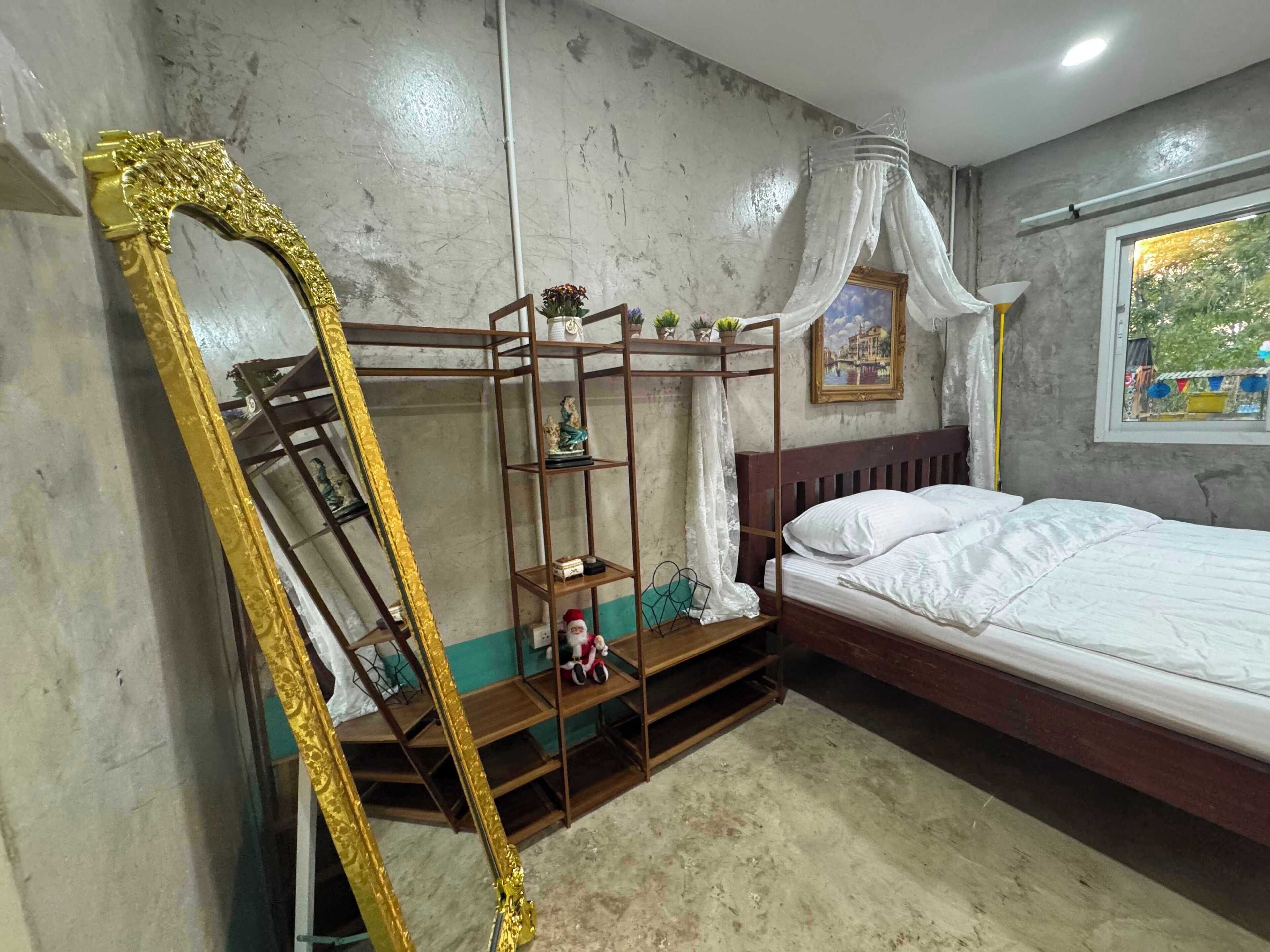 A bedroom with a bed, a wooden shelf with decorative items, a large mirror, and a window with a view of greenery.