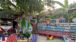 An outdoor seating area with wooden chairs and a table, surrounded by greenery and a decorative wall with multilingual text. - OYO 75539 Phanthida Resort and Cafe (Pranburi)
