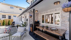A patio area with a wooden bench, a cushioned chair, and a small table. There is a white fence in the background, and a building with a brick facade is visible. - Beach Cottage 1-Block to Ocean! (Wildwood)