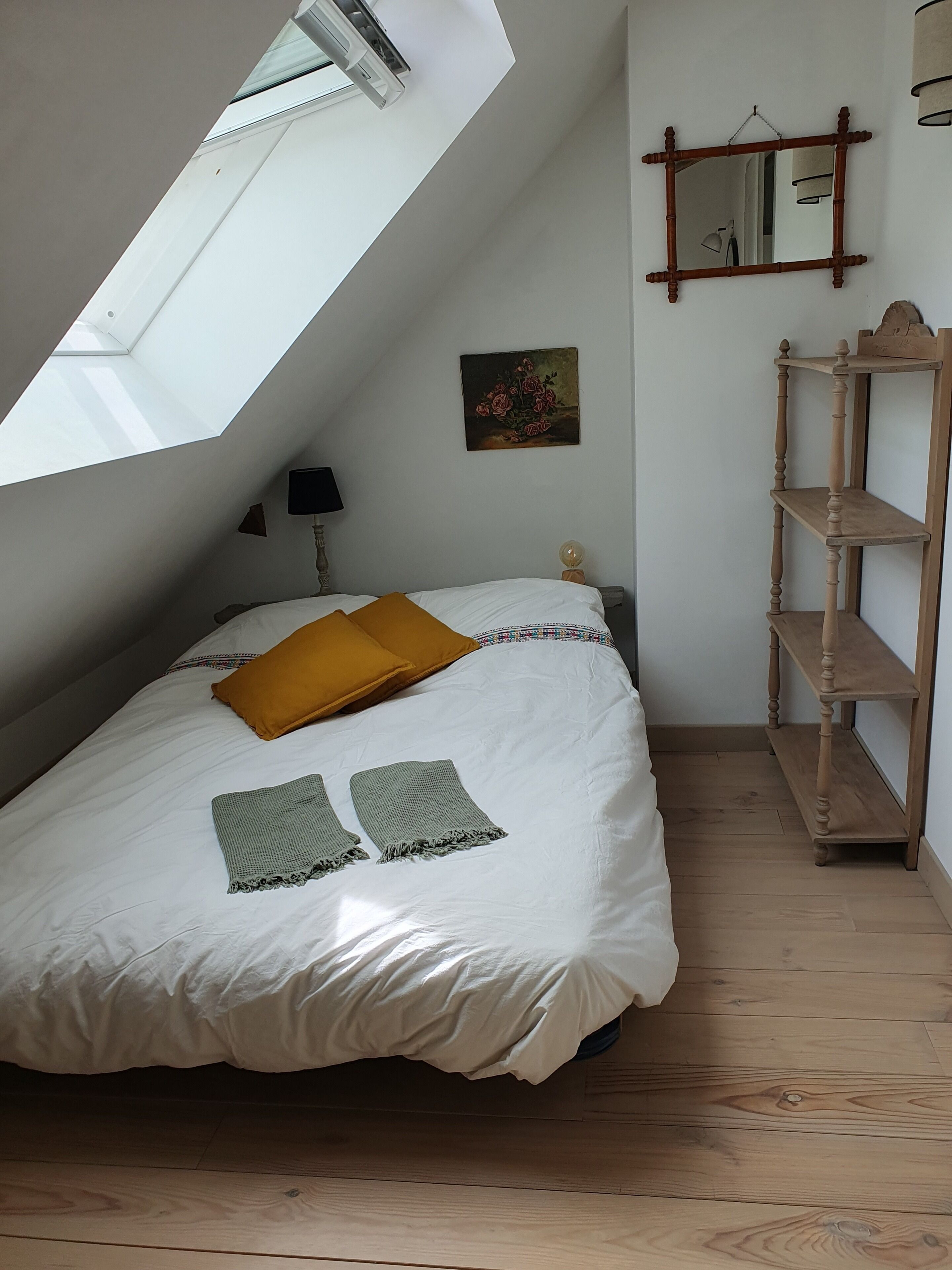 Ein Schlafzimmer mit schrägem Dach, einem Bett mit weißer Bettwäsche und einem senfkörnermitfarbenen Kopfkissen, einem kleinen Nachttischchen mit Lampe, einem wandmontierten Spiegel und einem Holzregal.