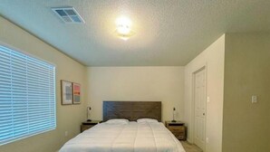Une chambre à coucher avec un grand lit, deux tables de chevet, une commode, un luminaire au plafond, une fenêtre avec des stores et une porte.