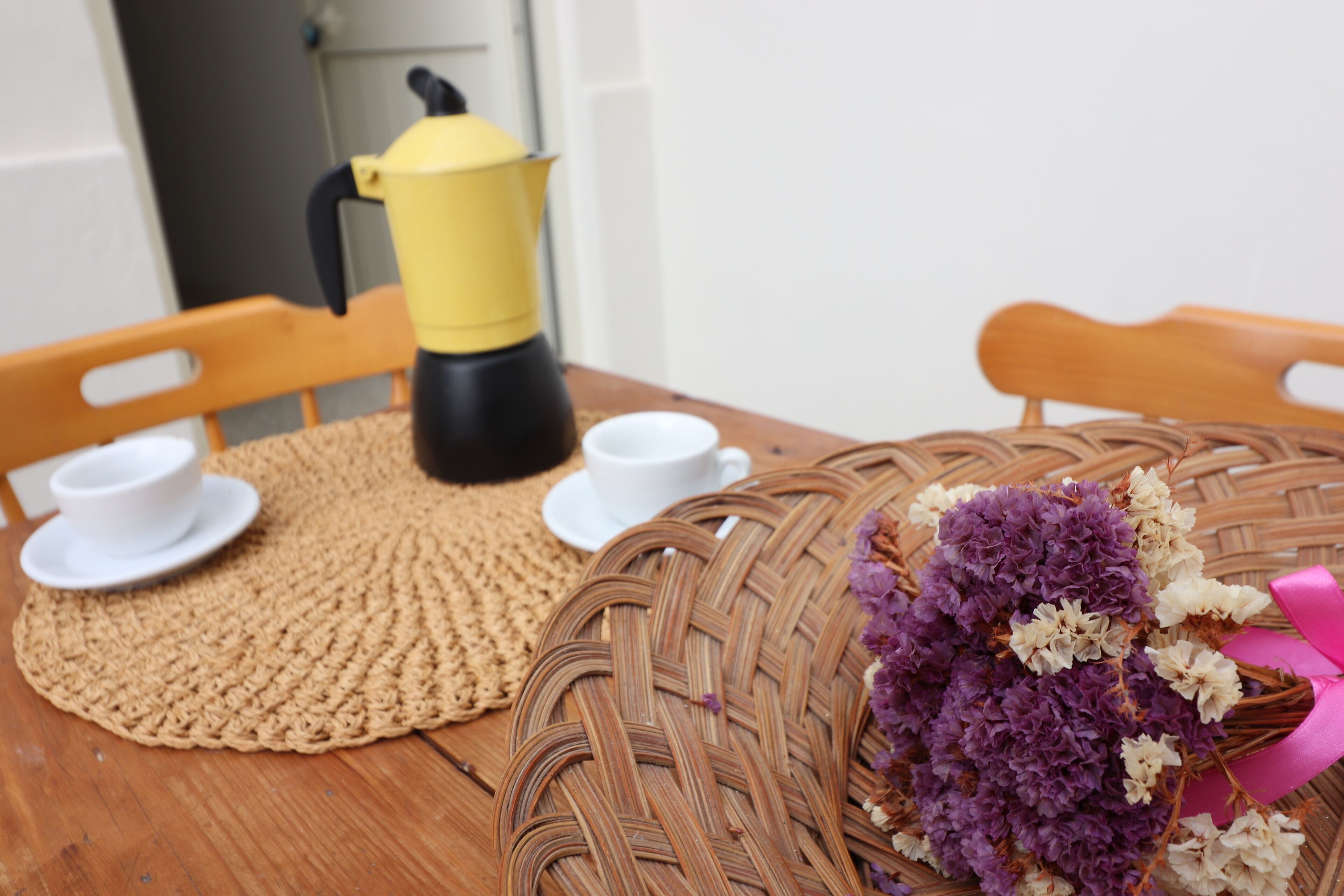 Et træbord med en vævet bordskåner, to hvide kopper, en gul kaffekande og et bundt lilla og hvide blomster.