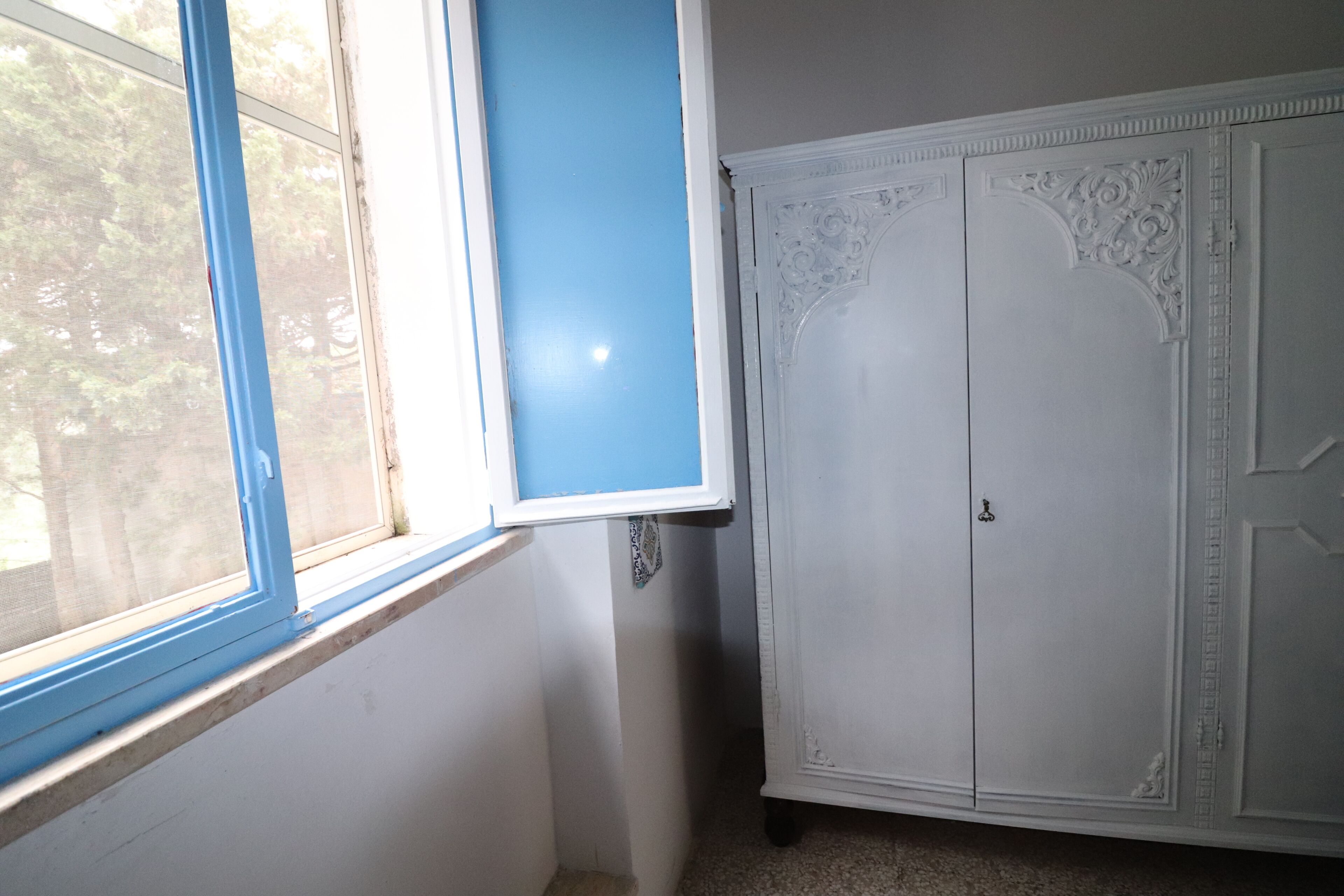 Ein Zimmer mit Fenster, ein weißer Schrank mit aufwendigen Verzierungen und einer blauen Wand.