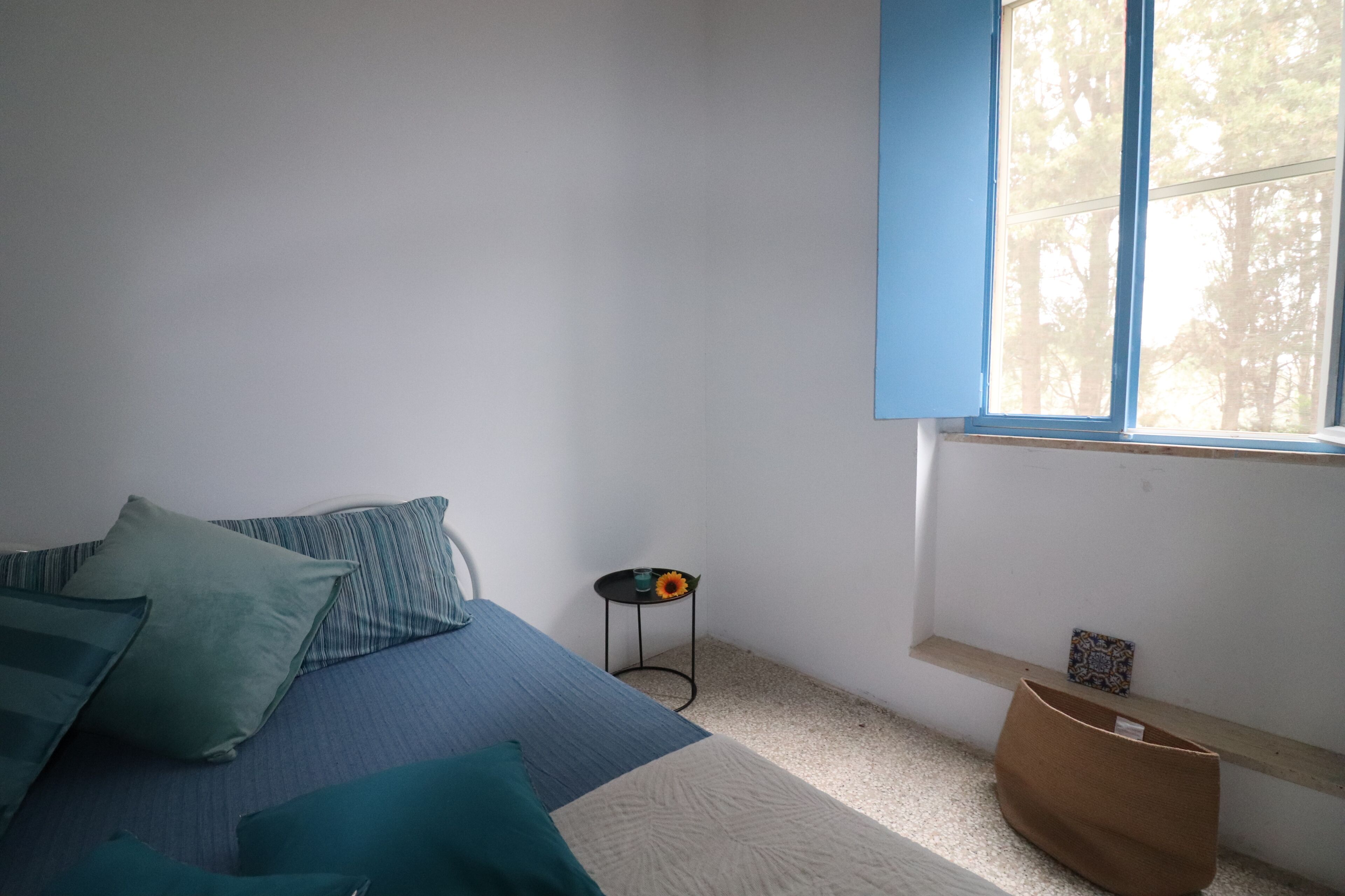 A bedroom with a bed, pillows, a small table with a flower, a window, and a shelf with a picture.