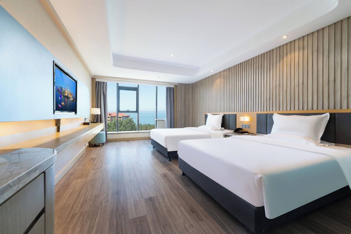 A modern hotel room with two beds, a large window with a city view, a flat-screen TV, and a wooden panel wall.