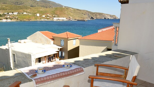 A balcony with a table set for two, overlooking a coastal view with houses and a clear sky. - Traditional island house (Andros)