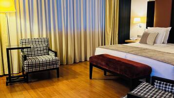 A hotel room with a bed, a chair, a bench, a lamp, and a curtain.