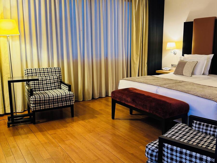 Quarto de hotel com cama, cadeira, banqueta, abajur e cortina.