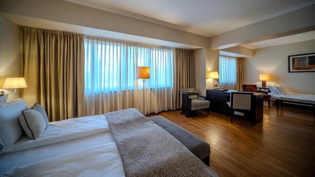A hotel room with a large bed, a desk with a chair, a sofa, and a window with curtains. - Hotel Rivo Timisoara (Timisoara)