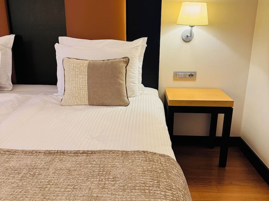 Una habitación de hotel con una cama grande, una mesita de noche, una lámpara y luz en la pared.
