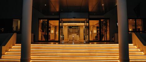 Entrada de hotel con fachada de vidrio, una escalera que conduce a la entrada y un camino bien iluminado.