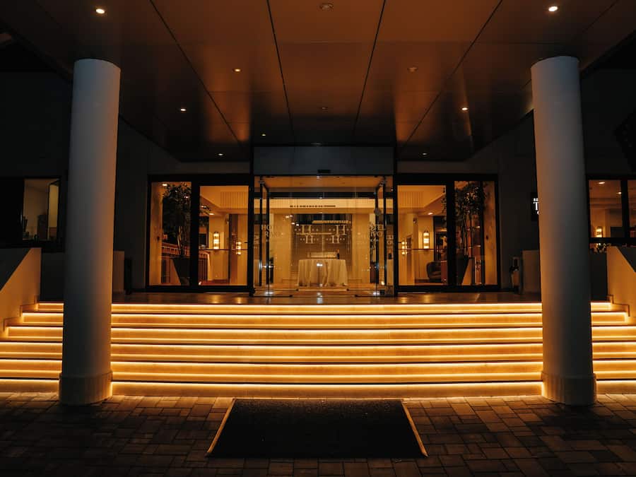 Entrada de hotel con fachada de vidrio, una escalera que conduce a la entrada y un camino bien iluminado.