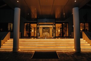 A hotel entrance with a glass facade, a staircase leading up to the entrance, and a well-lit pathway.