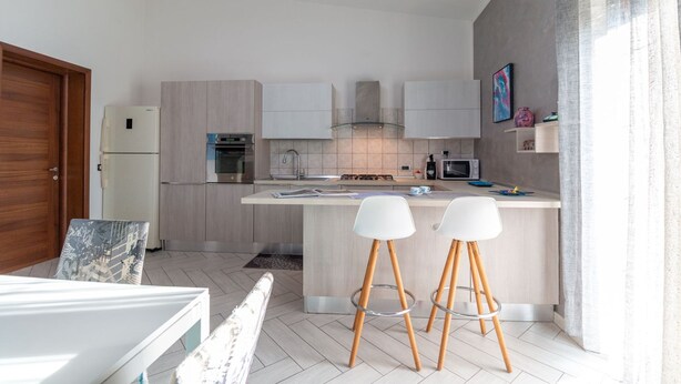 A modern kitchen with a central island, two bar stools, a refrigerator, an oven, a microwave, and a sink. - Mameli Apartment in Nurachi (Nurachi)