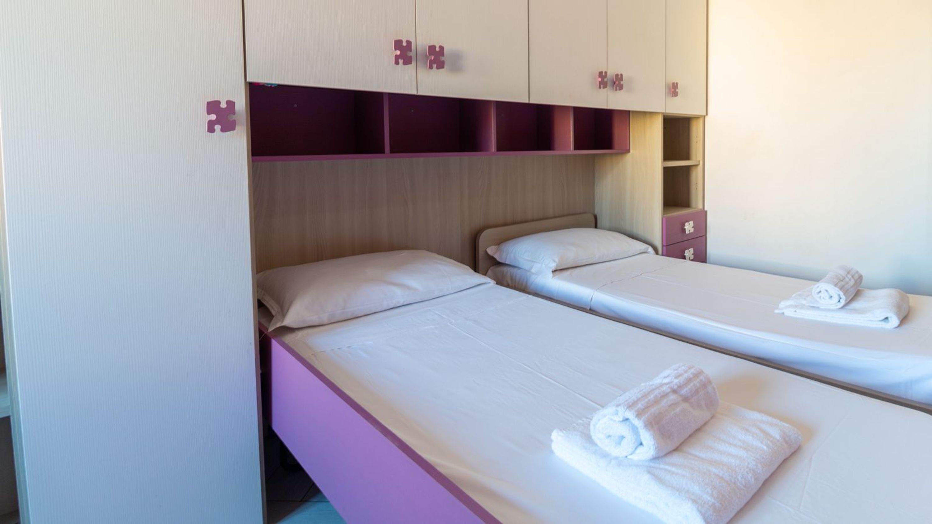 A room with two beds, each with a pillow and a rolled towel. There are cabinets above the beds with handles.