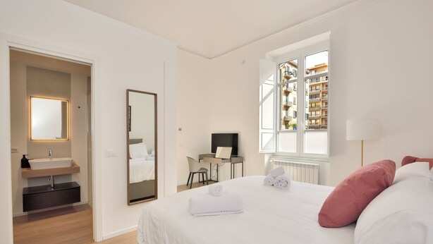 A hotel room with a bed, a desk, a chair, a TV, a window, and a bathroom with a sink and mirror. - Palazzo Gregorietti Boutique Apartments (Palermo)