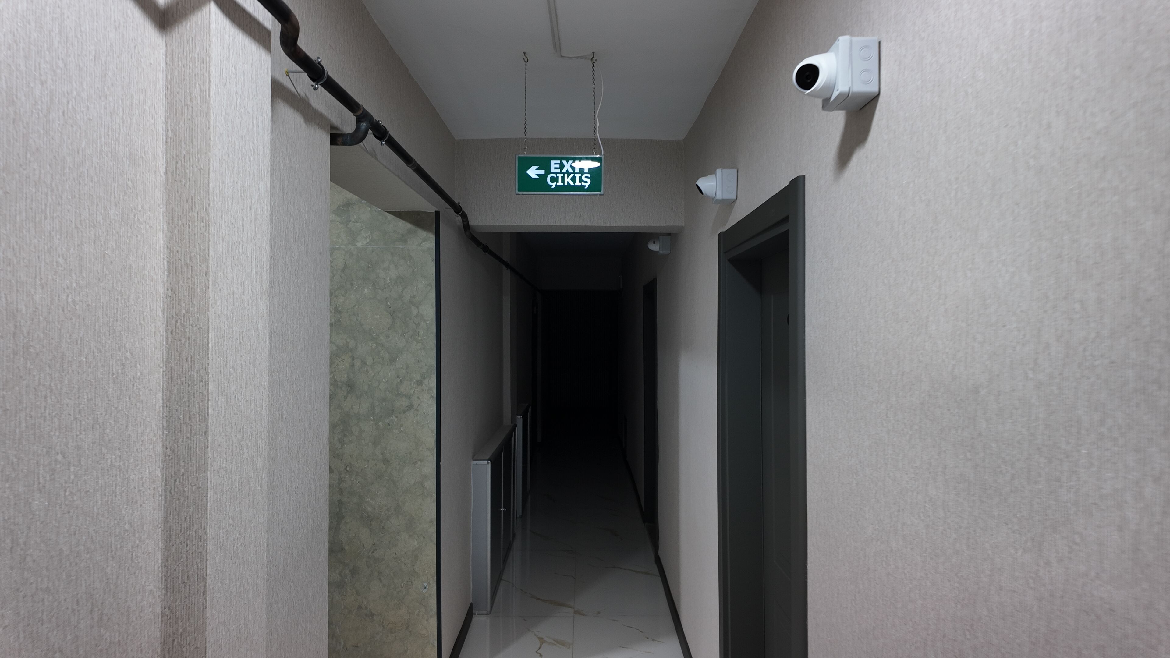 A narrow hallway with a green exit sign, a security camera, and a door on the right.