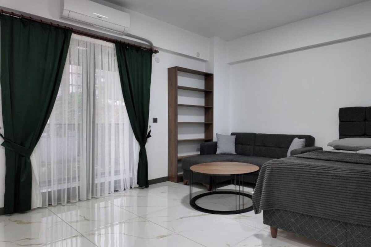 A modern hotel room with a bed, a sofa, a small round table, and a bookshelf.