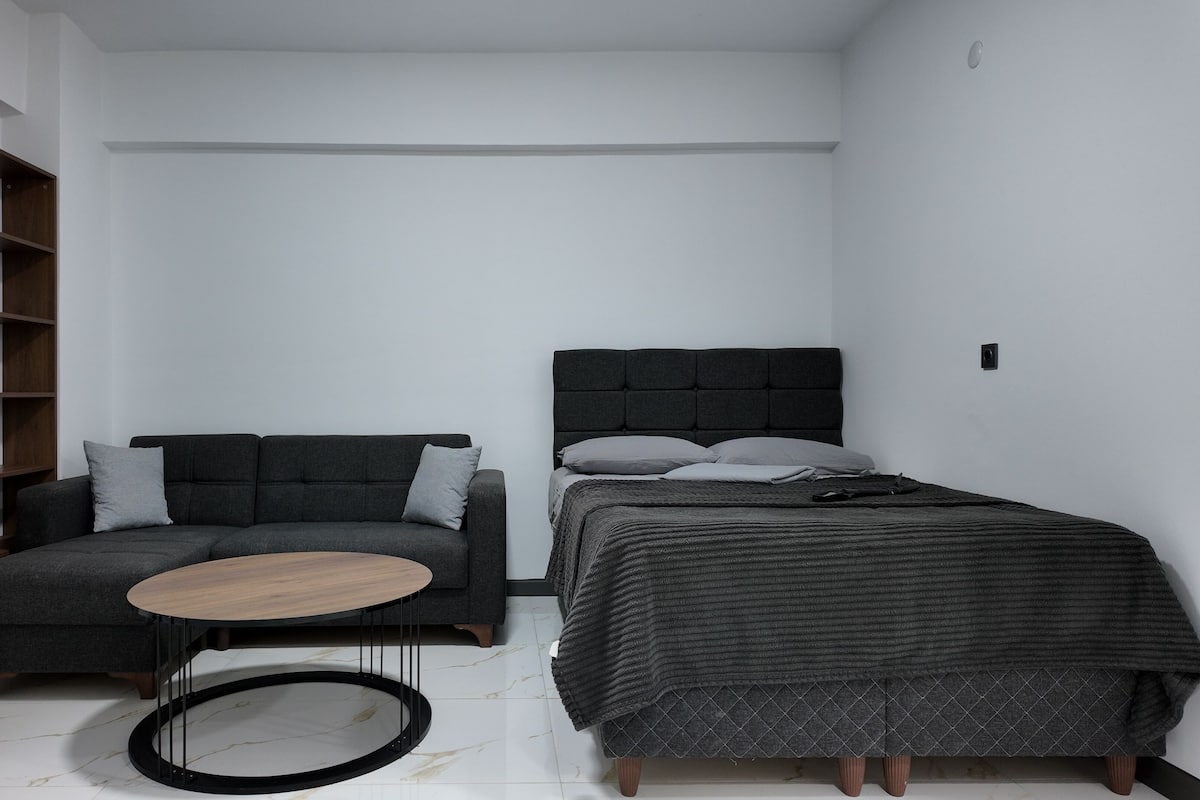 A small, modern room with a grey sofa, a round wooden table, and a bed with a grey headboard.